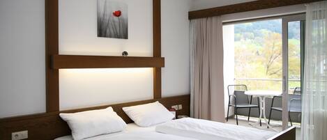 A hotel room with a large bed, a small table and chairs, a painting on the wall, and a balcony with a view of trees.
