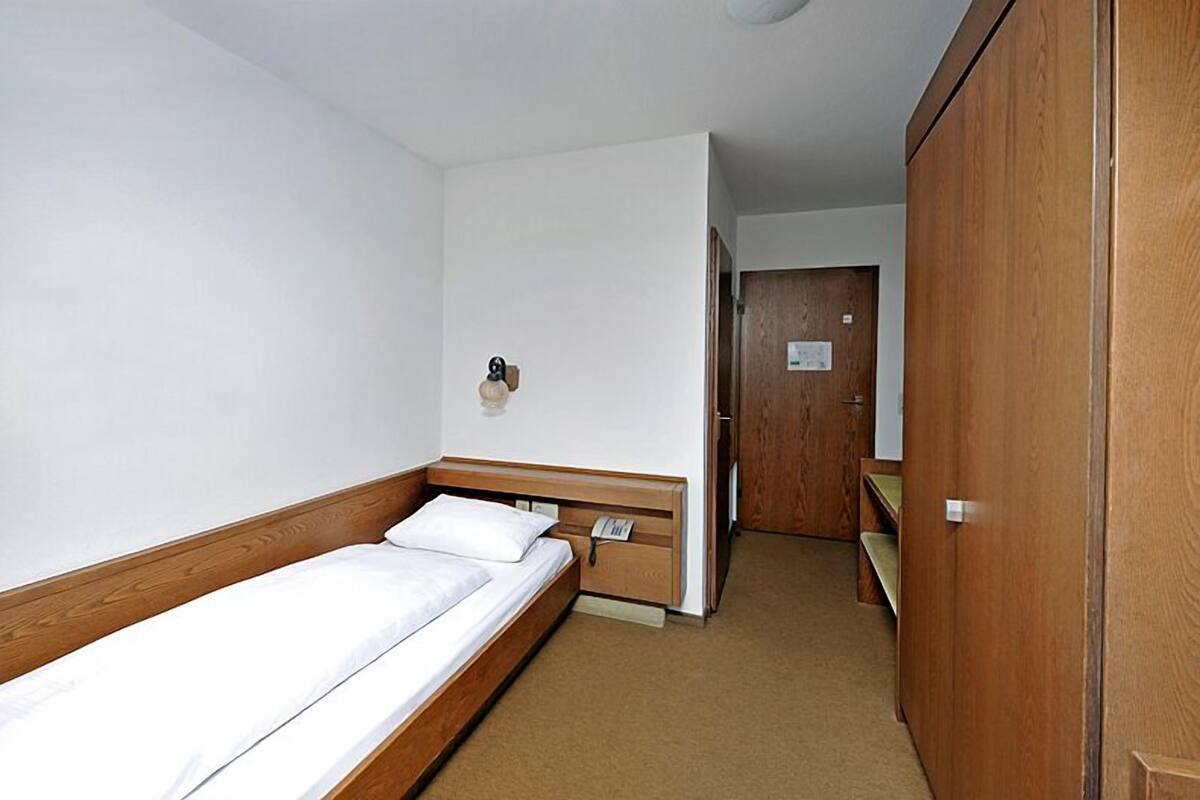 A small, single-bed room with a wooden headboard, a bedside table with a lamp, and a wooden wardrobe.