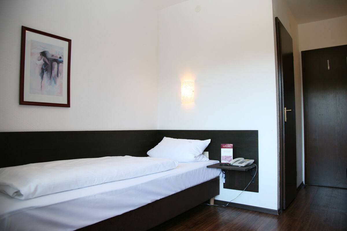 A single bed with white bedding, a bedside table with a phone, a framed picture on the wall, and a door to the right.