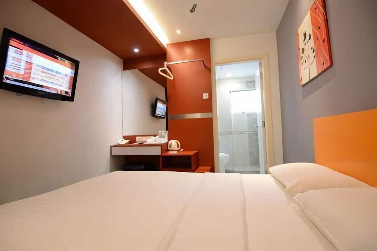 Quarto de hotel com cama, escrivaninha com computador, televisão e banheiro visível por uma porta aberta.