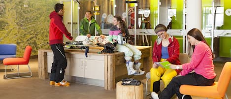 A group of people in a brightly lit indoor space with a wooden table, chairs, and a large wall mural.