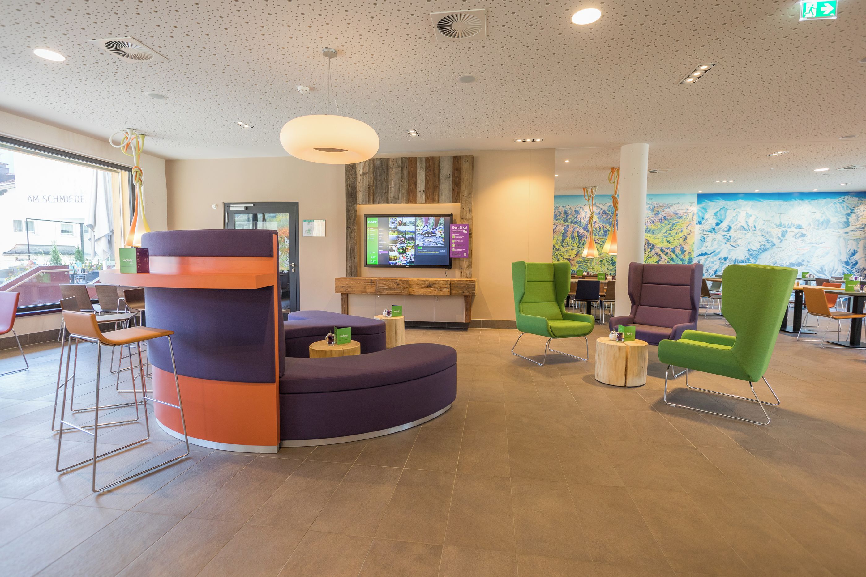A modern lobby with a curved seating area, a flat-screen TV mounted on a wooden wall, and a large mural depicting a mountainous landscape.