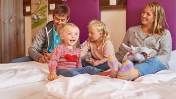 Eine Familie sitzt auf einem Bett mit einem violetten Kopfteil, einer Holzwandverkleidung und einer kleinen Pflanzendekoration.