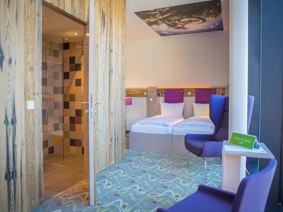 Habitación de hotel con ducha, cama con dos almohadas, una mesita y un cartel verde, además de una ventana con vistas al paisaje.