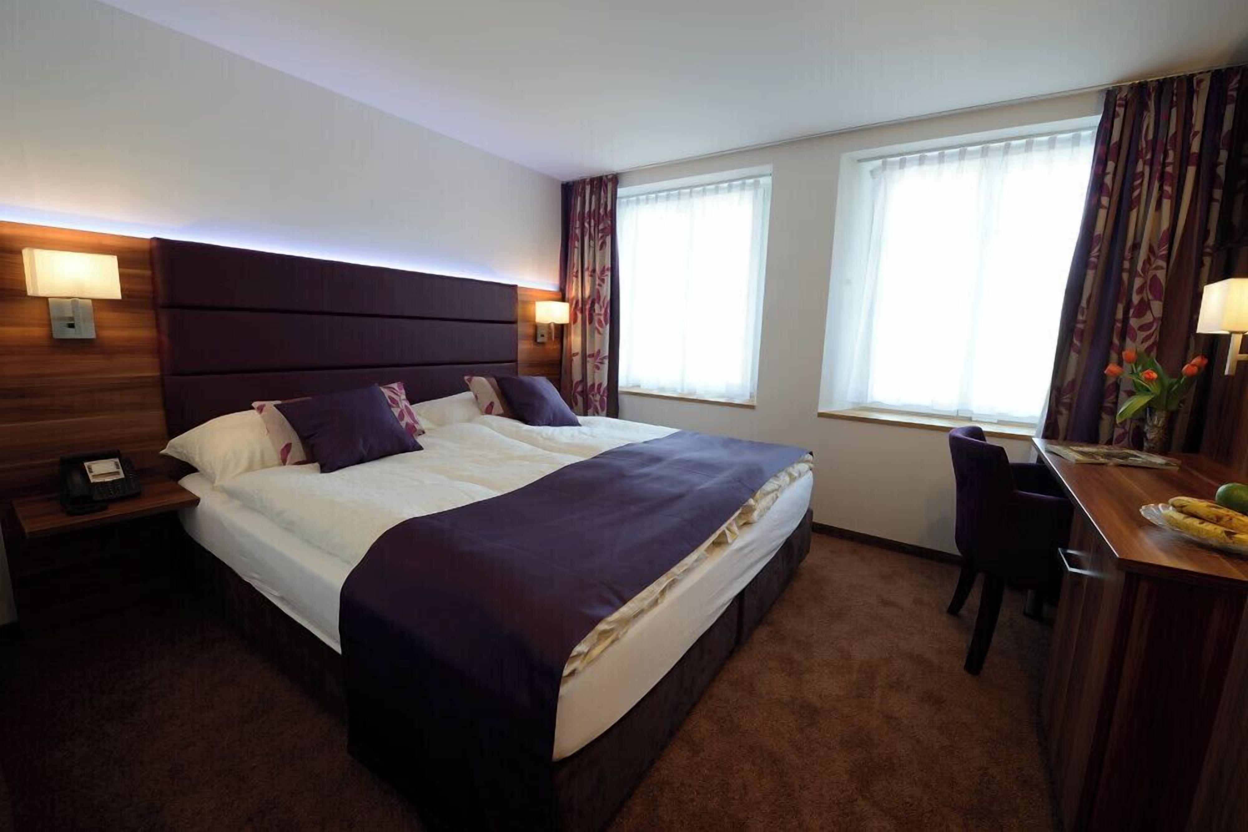 A hotel room with a large bed, two bedside lamps, a desk with a chair, and two windows with curtains.