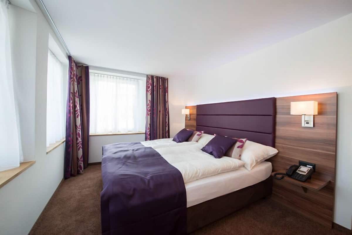 Quarto de hotel com uma cama grande, cabeceira de madeira, duas luminárias de cabeceira, um telefone e uma janela com cortinas.