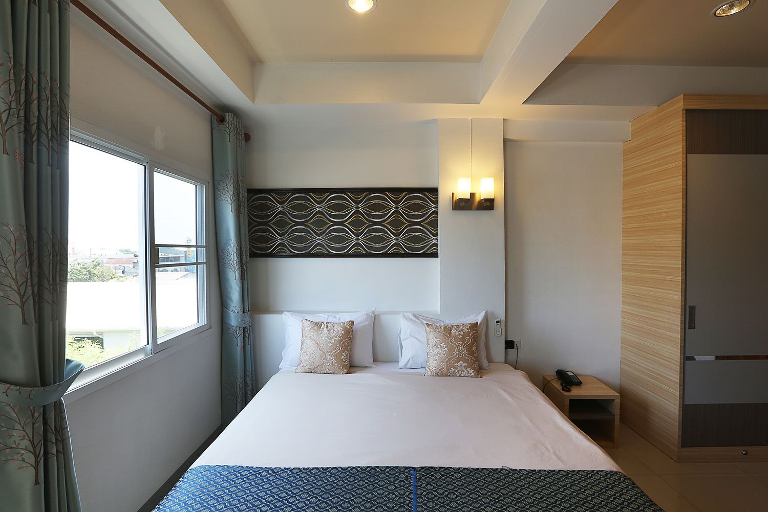 Habitación de hotel moderna con una cama grande, un cabecero estampado, una mesita de noche con lámpara y vistas al exterior.