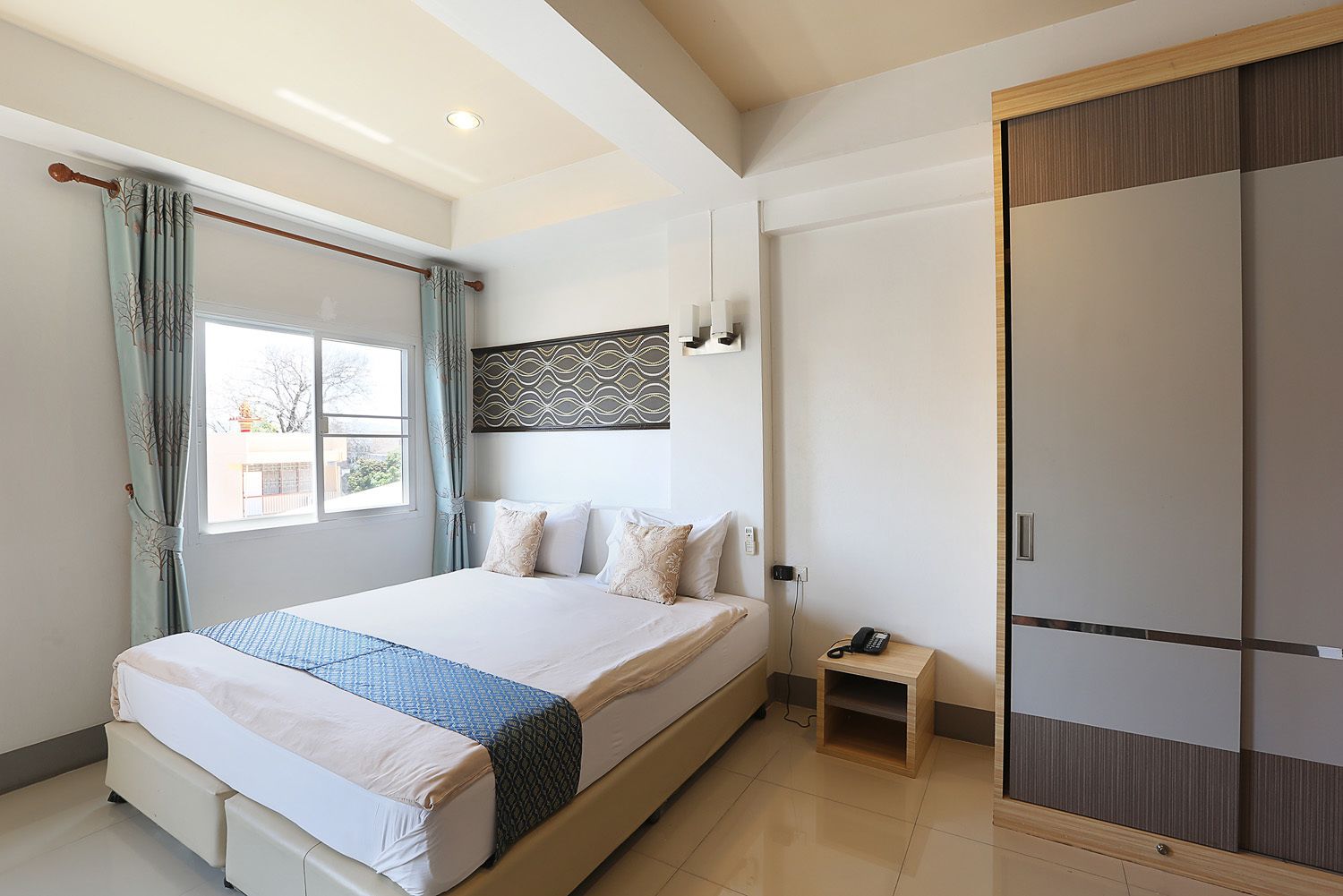A bedroom with a large bed, a bedside table with a phone, a wardrobe, and a window with curtains.