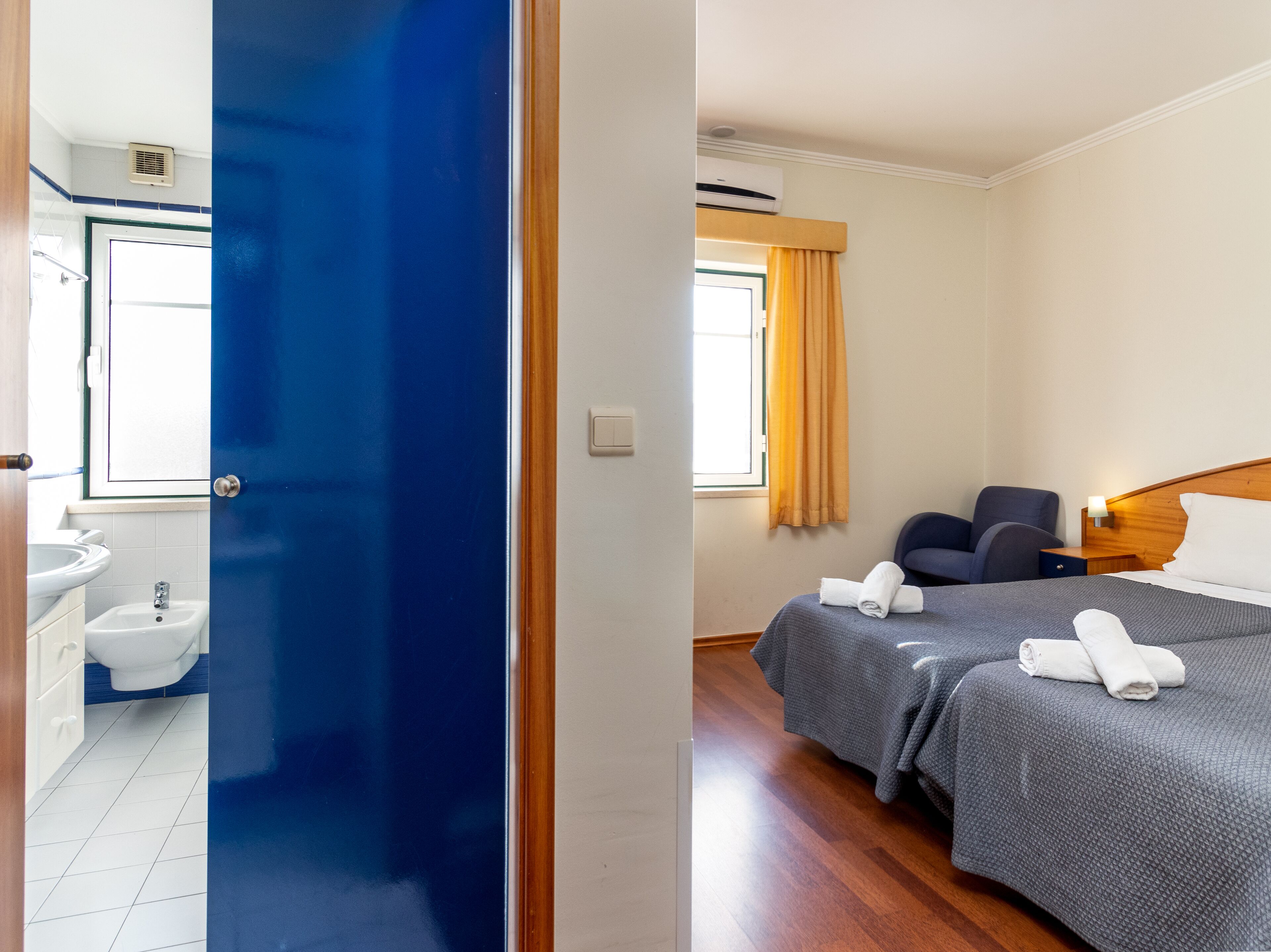 A hotel room with a bathroom, two beds, a bedside table, and a window with curtains.