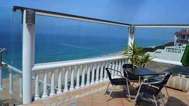 Un balcon doté d’une balustrade en verre donnant sur la mer, une table avec des chaises et une plante en pot.