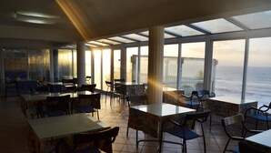 Un restaurant doté de grandes fenêtres donnant sur l’océan, avec un intérieur moderne, des sols carrelés et un mobilier simple.