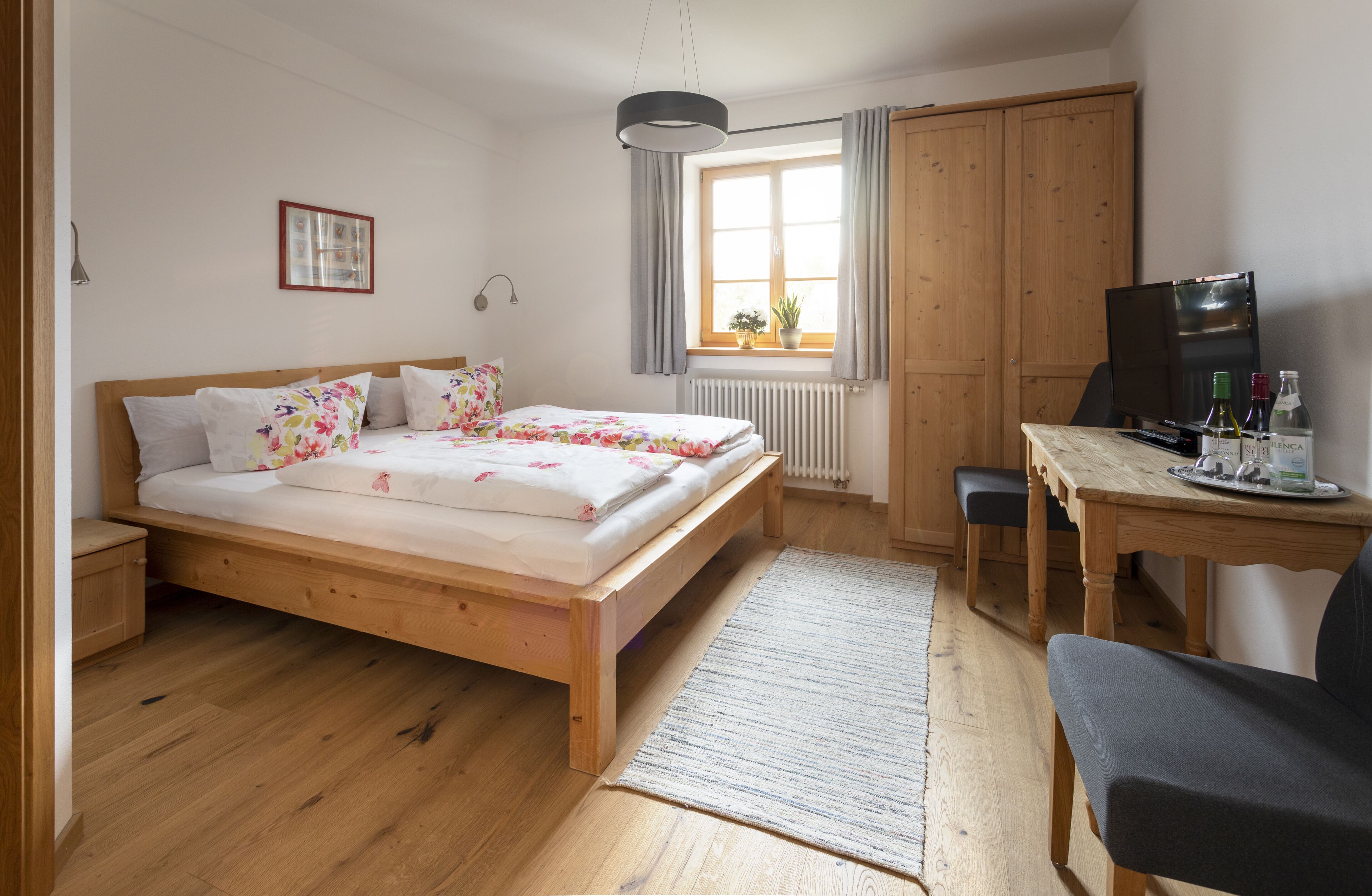 A bedroom with a wooden bed, a desk, a chair, a radiator, a window with curtains, and a television.