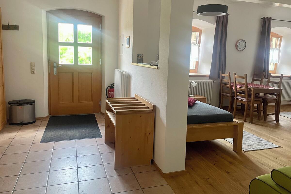 Una habitación con puerta de madera, un radiador, una cama, una mesa de comedor con sillas y un reloj en la pared.