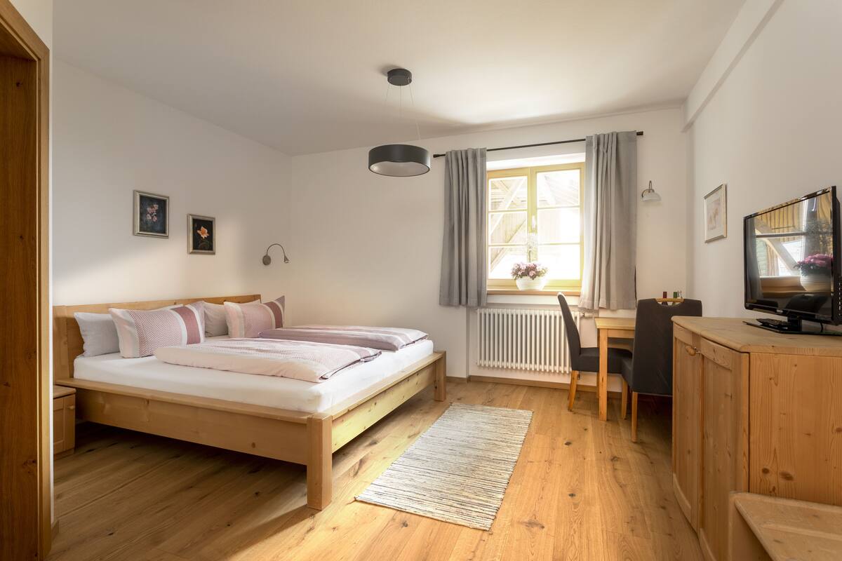 Un dormitorio con cama de madera, un escritorio, una silla, un televisor y una ventana con cortinas.