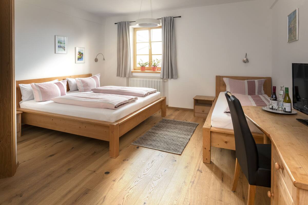 Un dormitorio con una cama de madera, un escritorio y una silla. Hay una ventana con cortinas, un radiador y una mesita con botellas.
