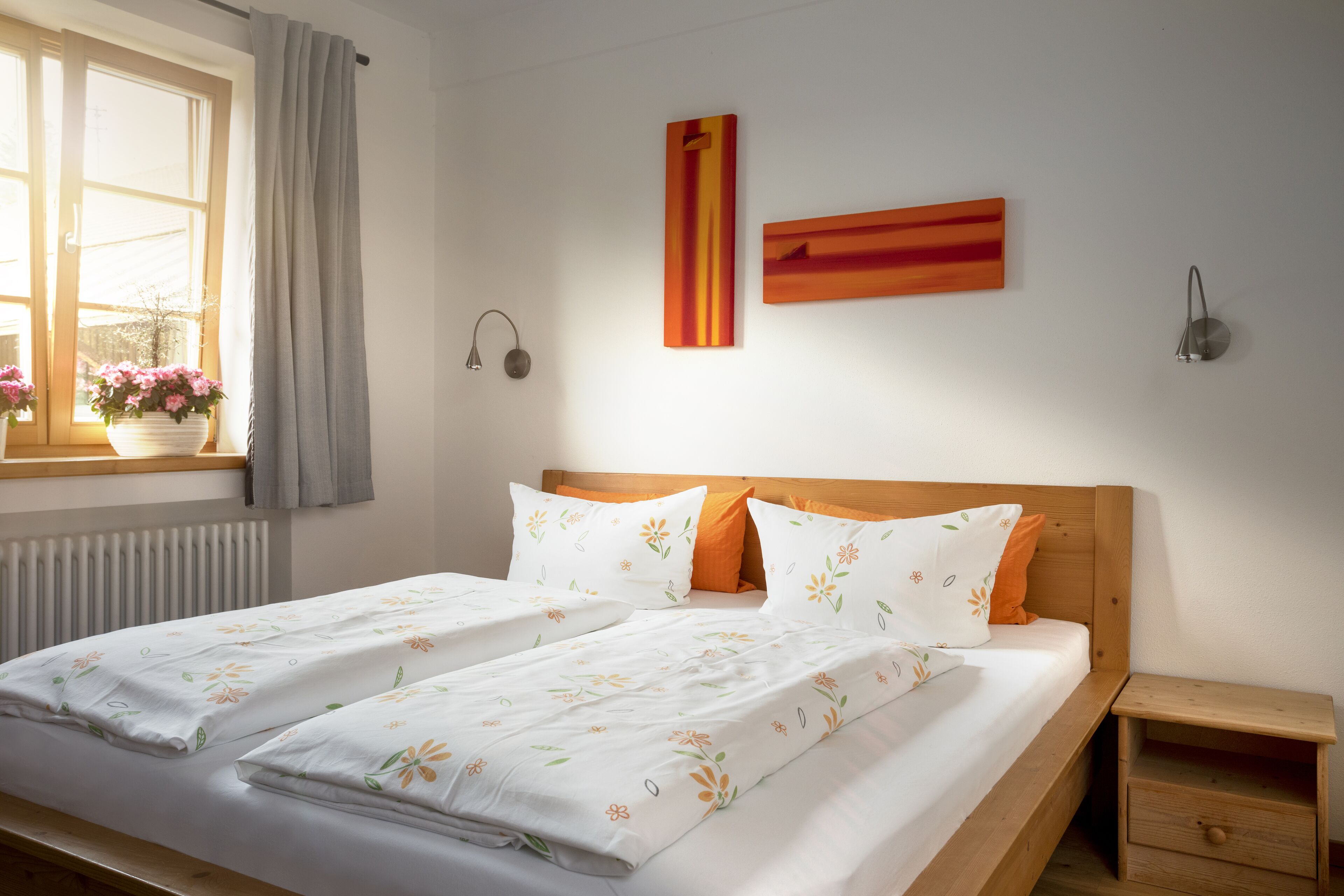 A double bed with floral bedding, a wooden headboard, a bedside table, and a window with curtains.
