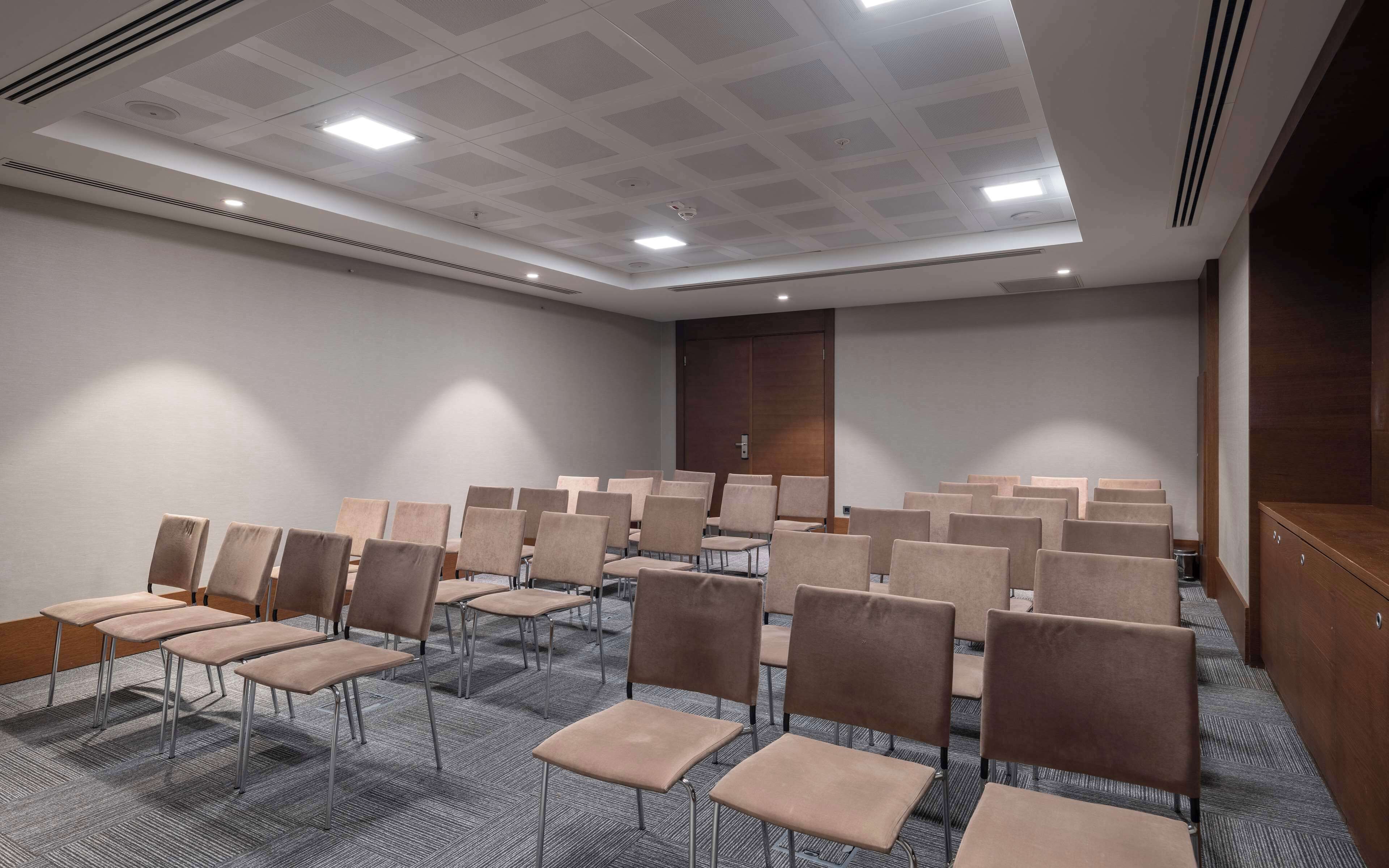 Een vergaderruimte met rijen stoelen, een vloerbedekking, een houten deur en een plafond met inbouwverlichting.