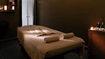 A spa room with a massage table, towels, candles, and toiletries on a wooden cabinet.