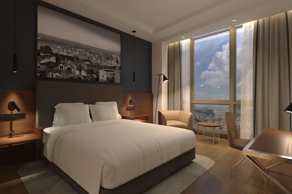 A modern hotel room with a large bed, a desk, a chair, and a city view through large windows.