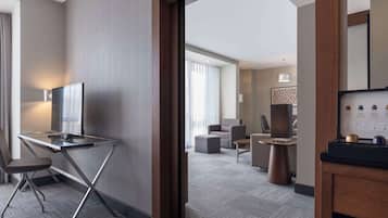 Una habitación de hotel moderna con escritorio, silla y televisor. Se observa un área de comedor con mesa y sillas al fondo.