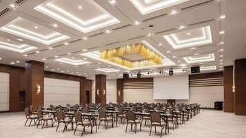 Una sala de conferencias con un diseño moderno en el techo, varias mesas con sillas y una pantalla grande para presentaciones.