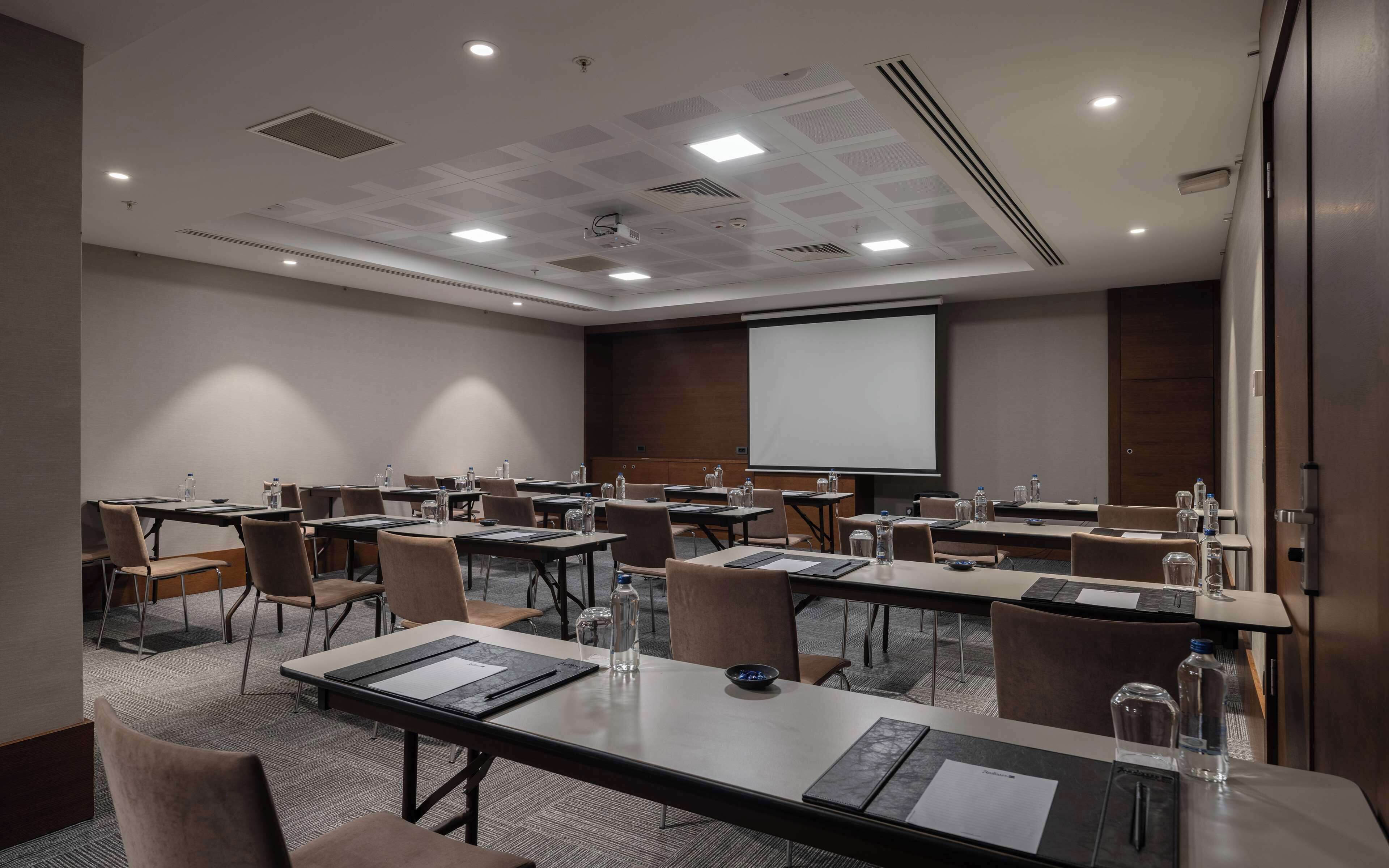 Un salón de conferencias con varias mesas, sillas, una pantalla de proyector y una pizarra.