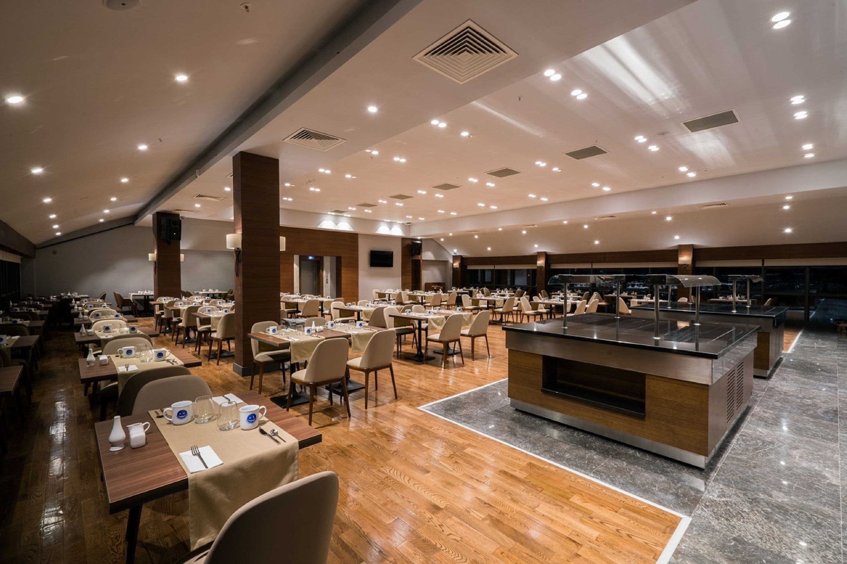 A spacious dining area with multiple tables and chairs, a central buffet station, and a high ceiling with recessed lighting.