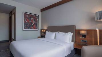 A hotel room with a large bed, a nightstand with a lamp, a wall-mounted TV, and a framed abstract painting.