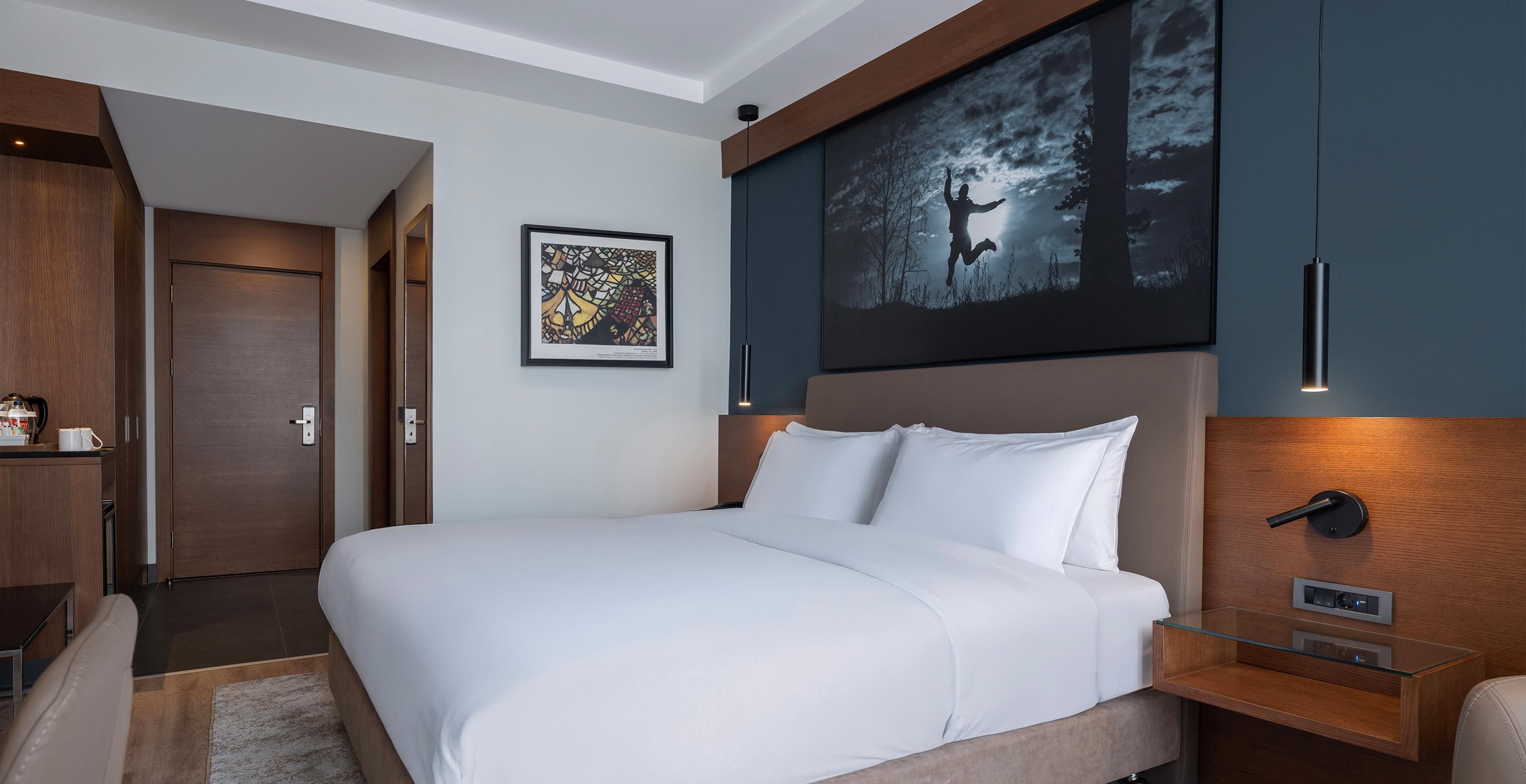 A modern hotel room with a large bed, a wooden headboard, a glass bedside table, a framed artwork, and a door leading to a bathroom.