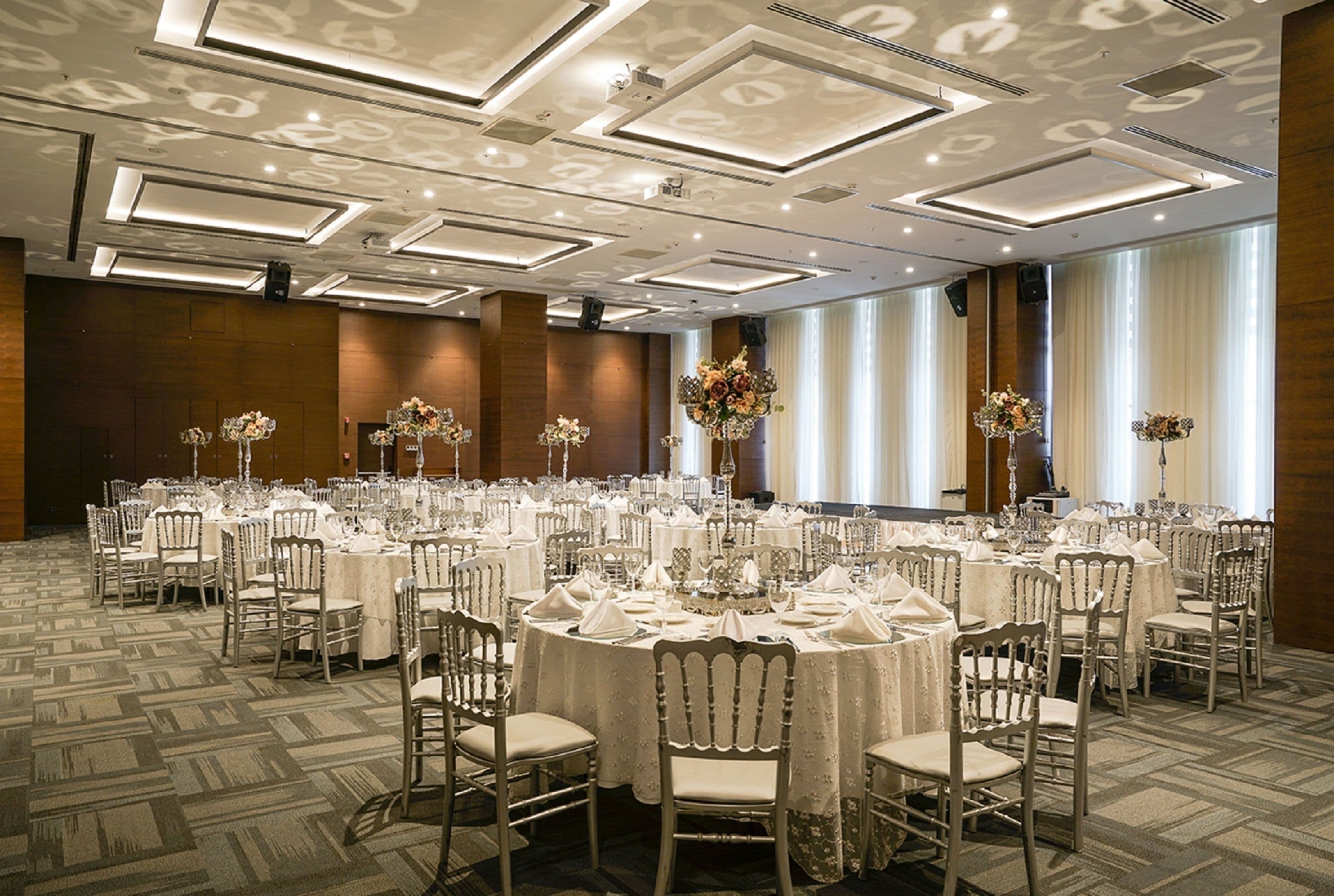 Un salón de banquetes con mesas redondas dispuestas para eventos, con alfombra estampada y un diseño moderno de techo.