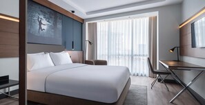 Habitación de hotel moderna con una cama grande, un escritorio, una silla, un televisor y un amplio ventanal con cortinas.