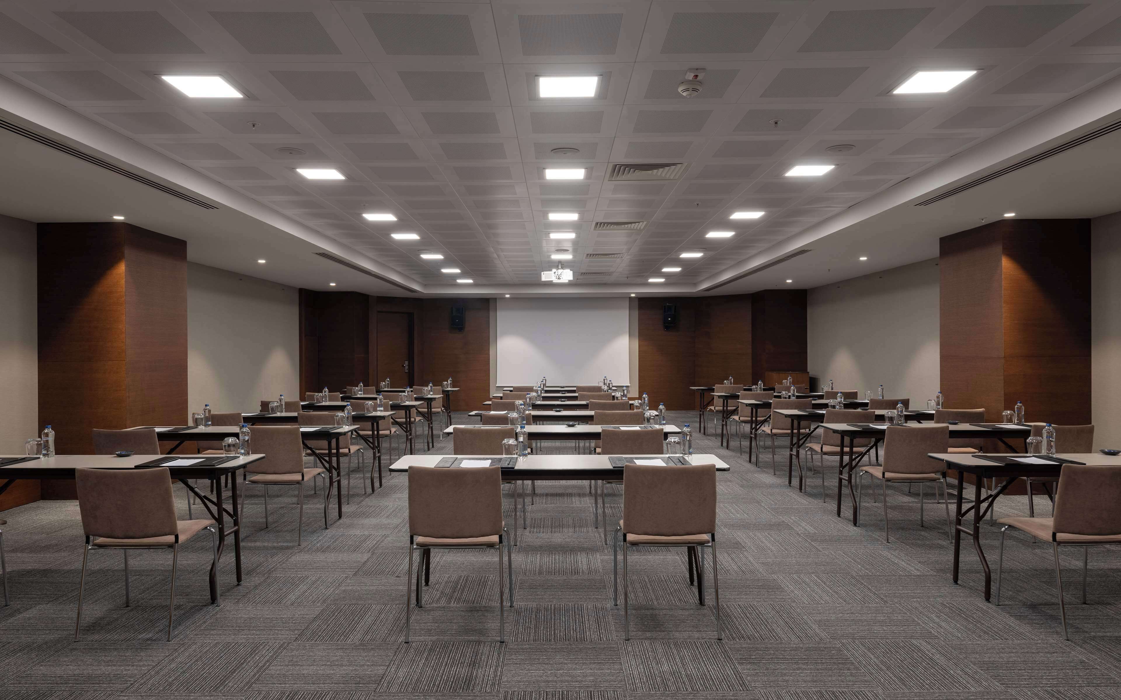 A conference room with multiple tables, chairs, and a large screen at the front.