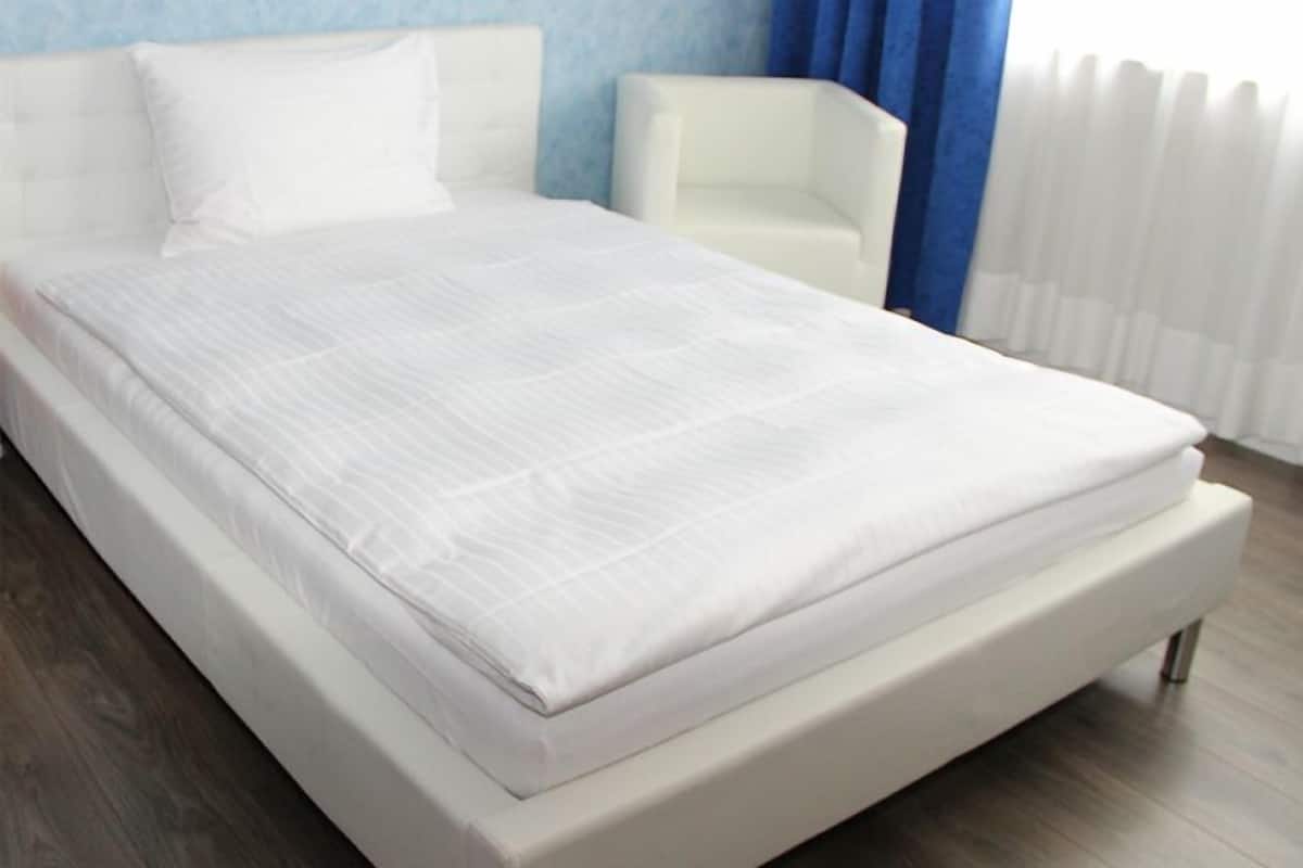 Cama bem arrumada com roupa de cama branca em quarto com parede azul e cadeira branca.
