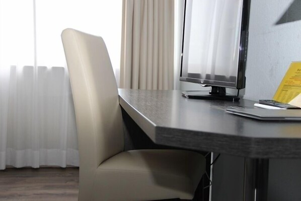 A modern office chair, a desk, and a computer monitor in a room with a window and curtains. - Parkhotel David (Loerrach)