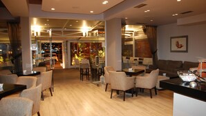 A modern hotel lobby with a wooden floor, seating areas, and a view of the city through large windows. - Hotel Mano (Sarandë)