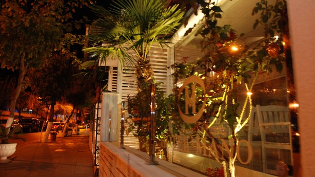 A nighttime street view with a building featuring a large illuminated 'M' sign, a palm tree, and a brick wall. - Hotel Mano (Sarandë)