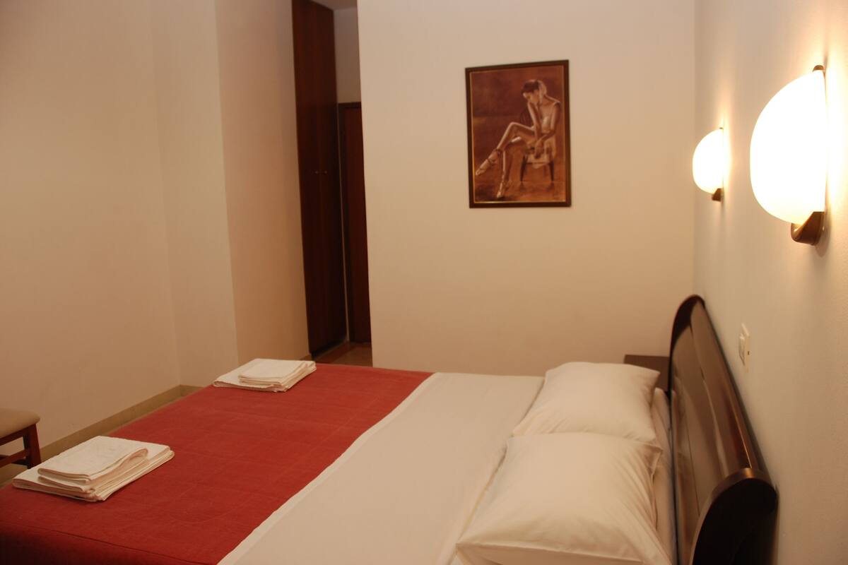 Une chambre d’hôtel avec un lit, une couvre-lit rouge, des oreillers blancs, une tête de lit en bois, un tableau au mur et une table de chevet avec une lampe.