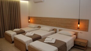 A hotel room with four single beds, each with a wooden headboard, white linens, and a brown throw blanket. There is a wooden nightstand with a drawer, a wall-mounted air conditioner, and a pendant light above each bed. - Hotel Mano (Sarandë)