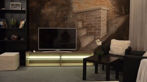 A modern living room with a flat-screen TV on a wooden stand, a black sofa, a white fluffy pillow, and a small table with a potted plant. - Guesthouse Soul Ljubljana (Ljubljana)