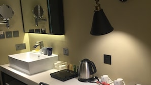 Habitación de hotel con un escritorio blanco, una silla negra, un lavamanos blanco, un espejo, una lámpara negra, una tetera y un plato con aperitivos.