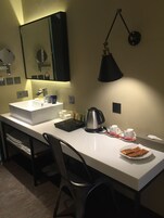 Habitación de hotel con un escritorio blanco, una silla negra, un lavamanos blanco, un espejo, una lámpara negra, una tetera y un plato con aperitivos.