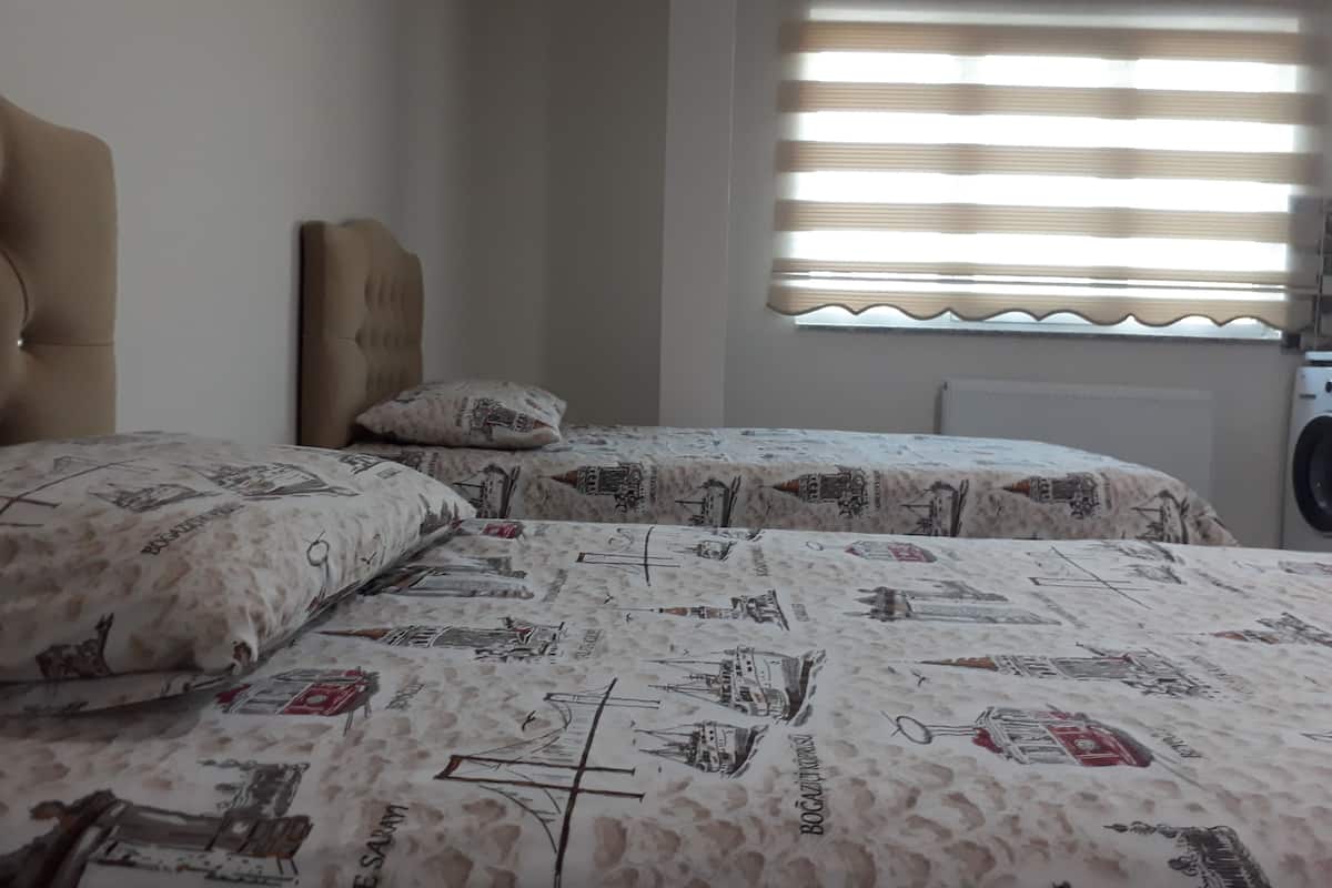 A bedroom with two beds, a headboard, a window with blinds, and a washing machine in the corner.