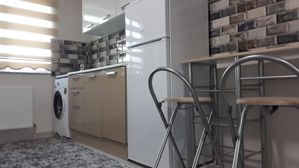 A compact kitchen with a washing machine, a refrigerator, and a metal bar stool. - Moonlight (Erzincan)
