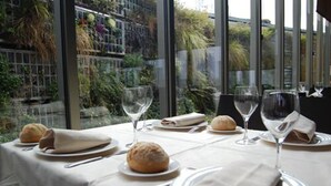 Una mesa de comedor dispuesta con platos, servilletas, pan y copas de vino, con vistas a la vegetación a través de amplios ventanales.