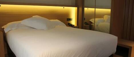 A neatly made bed with white linens, a bedside table with a lamp, and a mirror reflecting the room.