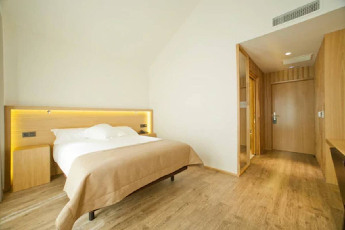 Una habitación de hotel moderna con piso de madera, una cama con cubrecama color beige, una mesita de noche y un guardarropa.