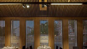 A restaurant with a long table set for dining, wooden slatted ceiling, and large windows with curtains.