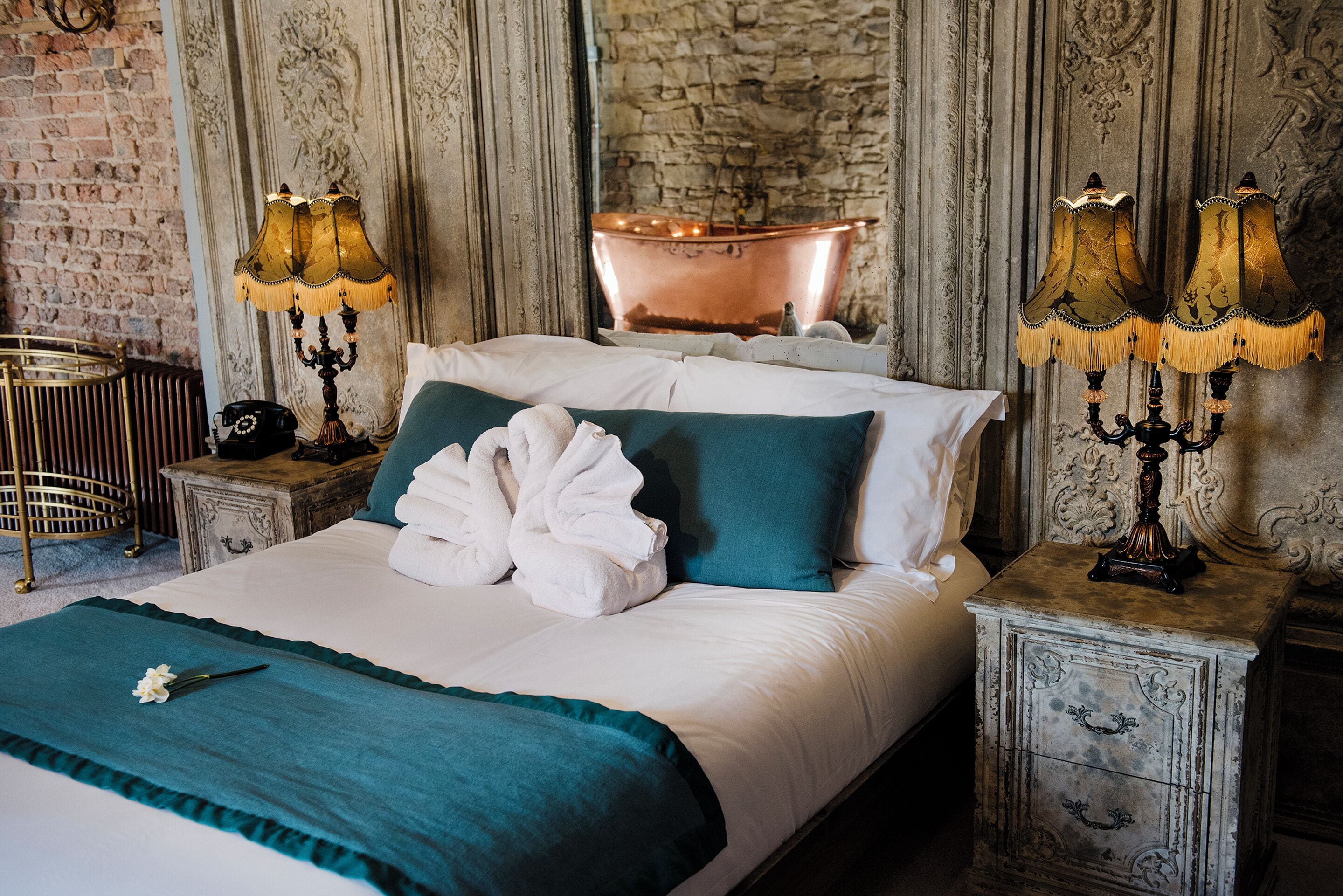 A neatly made bed with a teal throw blanket, white pillows, and a pair of white bathrobes. There are two wall-mounted lamps with fringed shades on either side of the bed. A copper bathtub is visible in the background, and a stone wall adds to the room's rustic charm.