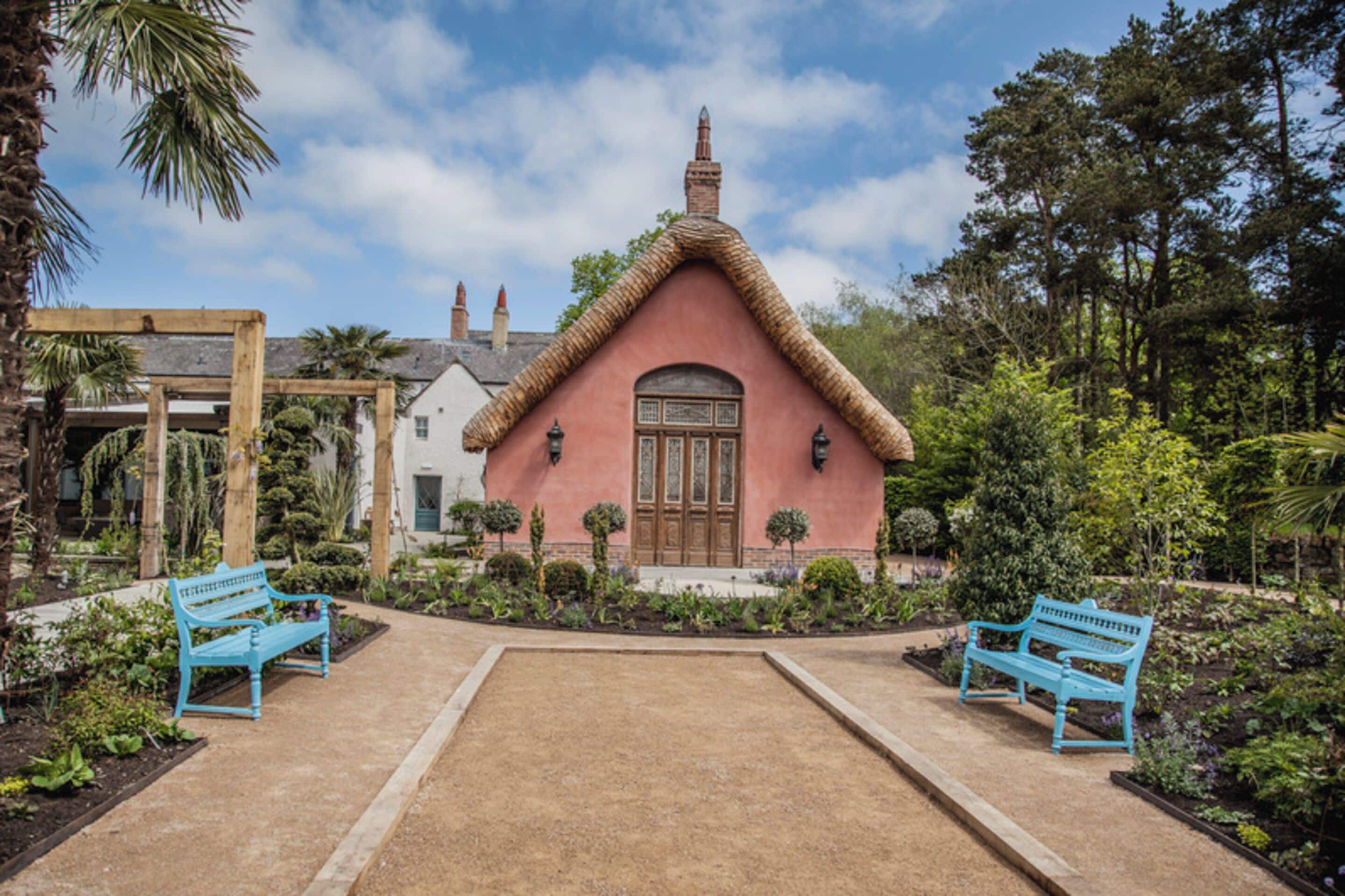 A pink building with a thatched roof, surrounded by a garden with benches.