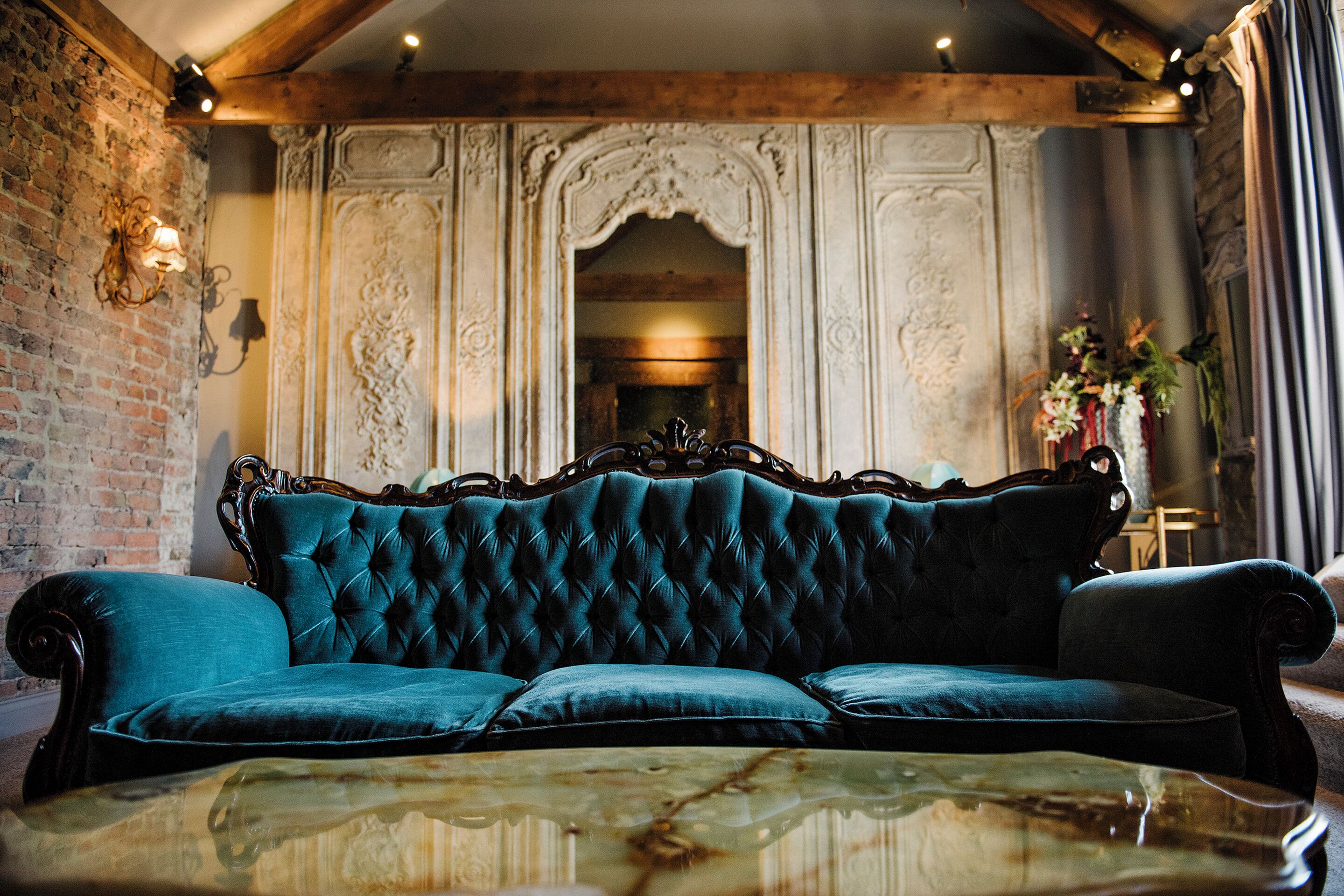 A luxurious teal upholstered sofa with ornate detailing, positioned in front of a decorative wall with intricate carvings and a mirror.